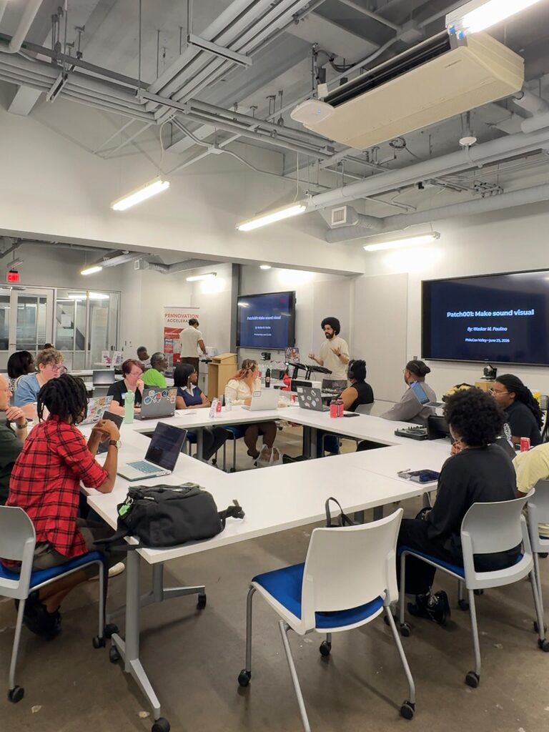 A wide view of the seminar room: people at laptops around U-shaped tables, a presenter at the front, and the PATCH 001 title on the screens.